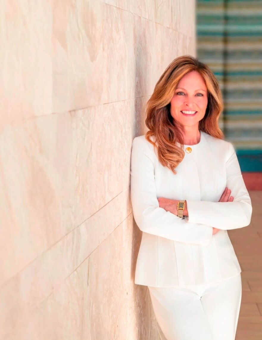 Blonde woman in white suit smiles with arms crossed next to wall 