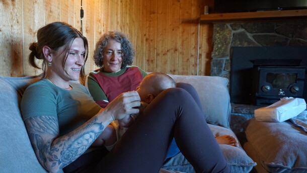  A woman with tattoos on her right arm holds a small baby in her lap on a grey couch. Another woman sits next to her, gazing at the baby.