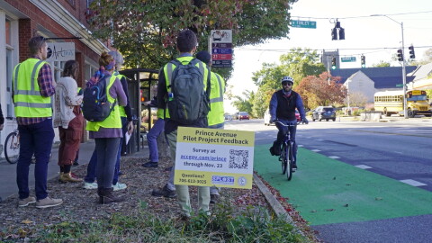 A bicyclist rides in a green bike lane along Prince Avenue while several people in safety vests stand beside a sign requesting feedback on a pilot project.