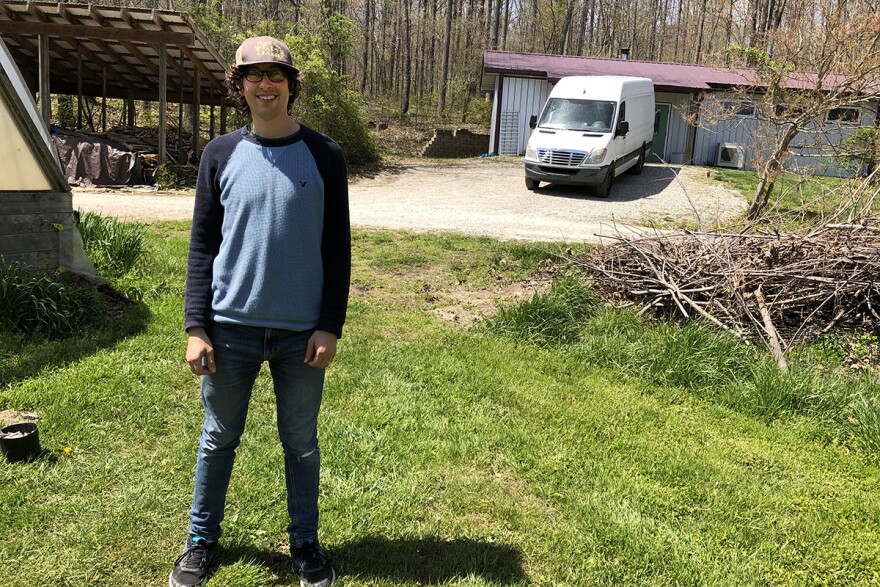 Eric Schedler, co-owner and head baker at Muddy Fork Bakery stands next to his greenhouse with their woodpile, delivery van, and bakery in the background.