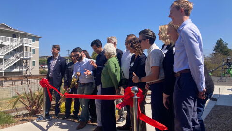 A person wearing a button-down shirt and sunglasses cuts a red ribbon alongside several other people.