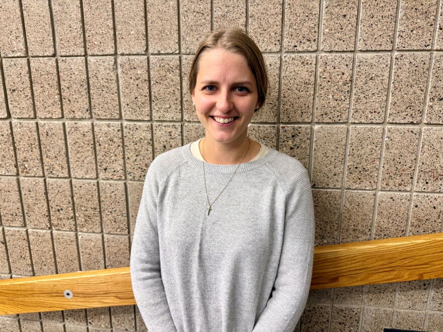 A woman who is smiling and wearing a grey sweater is standing next to a wall.