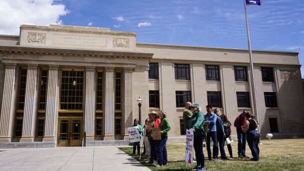 People stand with signs outside a tall cream building with pillars, amid a blue sky.