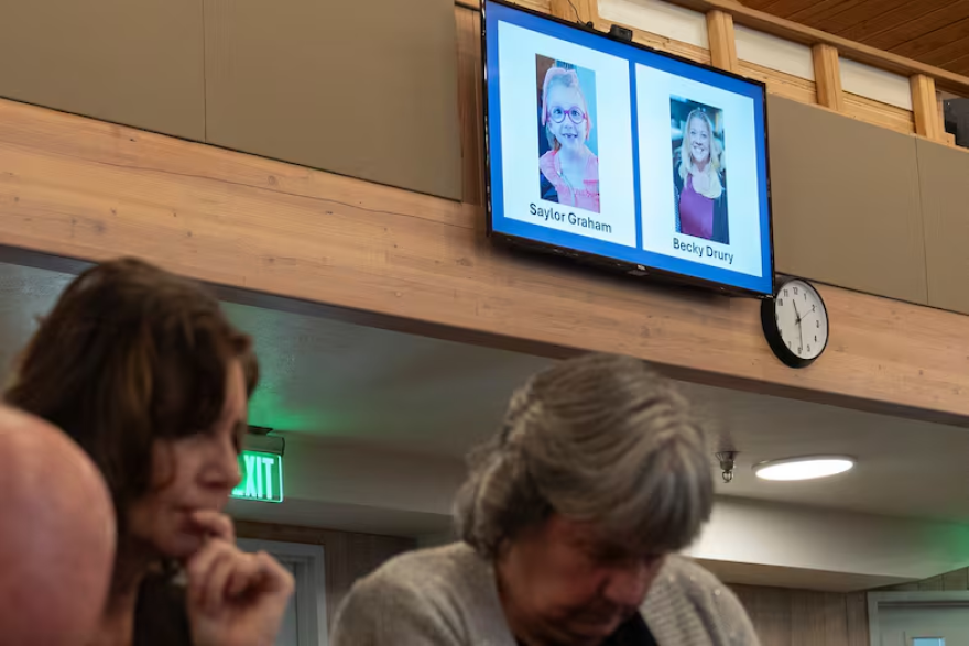 Two people bow their heads. On the wall behind them images of Saylor Graham, 7, and Becky Drury are displayed on a screen.