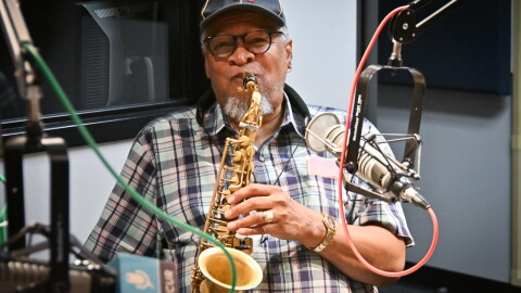 A man sits inside a radio studio playing a saxophone in front of a microphone.