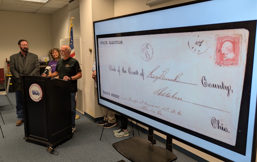 Three people stand at a lectern, while a historical document is displayed on a tv screen.