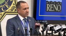 Corey Solferino standing behind a podium with mics pointed at him. The City of Reno logo is next to him on a TV screen.