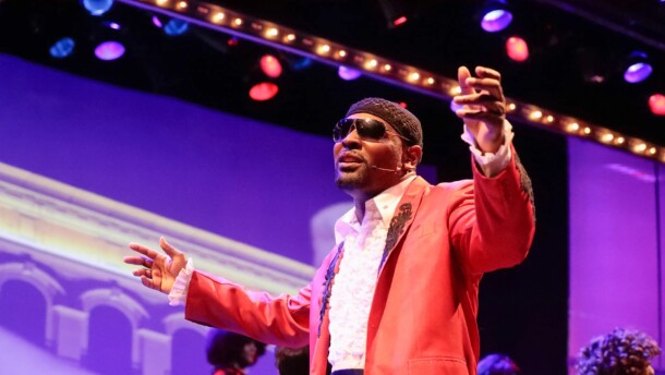 Black man wearing sunglasses and peach suit jacket singing on stage with his arms out