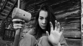 A woman sitting on the ground near the outside wall of a wooden structure holds a wooden mallet in one hand with her other hand raised in surrender.