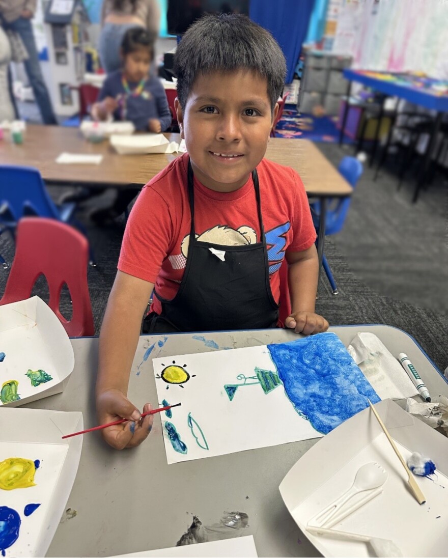 A young boy smiles for the camera as he sits at a table. He holds a paintbrush and is working on a painting of a boat on the water.