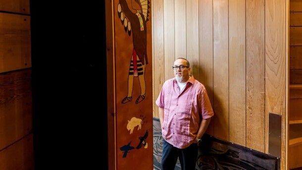 A man stands against a wooden wall and looks at the camera. One panel has a painting of a Native person in regalia. 