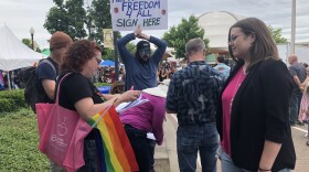 Profile view of a woman with pink tote bag and Pride flag, holding a petition on a clipboard, facing a woman who's wearing a dark jacket.