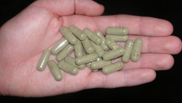A left hand holds a small pile of pill capsules filled with a green powder against a black backdrop.