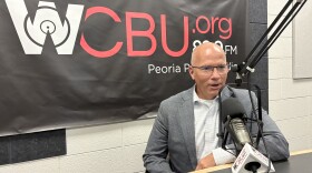 A man in a gray blazer and white shirt speaks into a microphone in a radio studio with a WCBU.org 89.9 FM banner behind him.