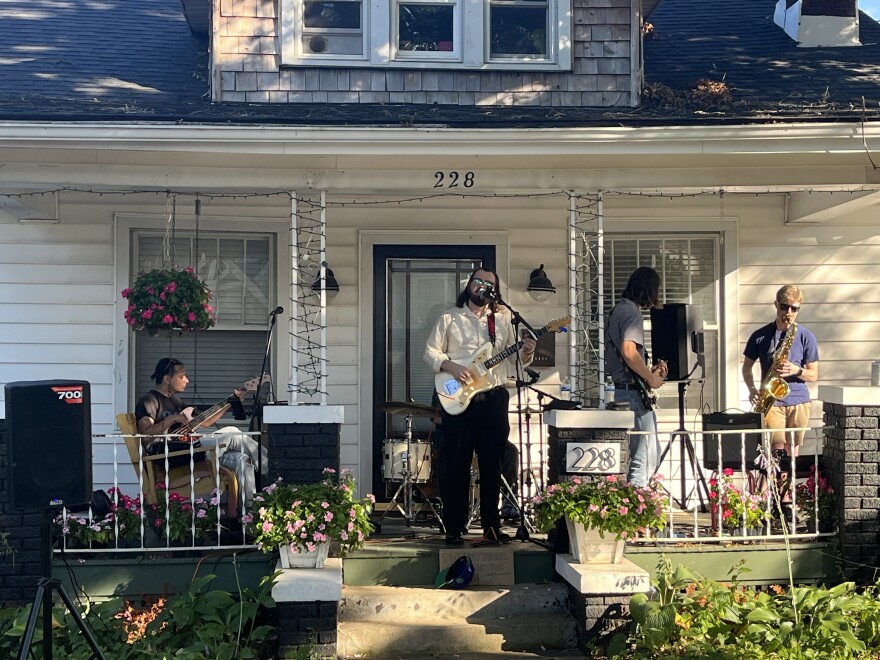 A band with two electric guitars, a bass, and a saxophone play on a front porch, facing out at the lawn.