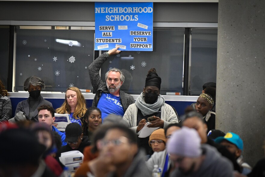 A man wearing a blue T-shirt holds a blue sign that reads "Neighborhood Schools Serve all students; deserve your support."