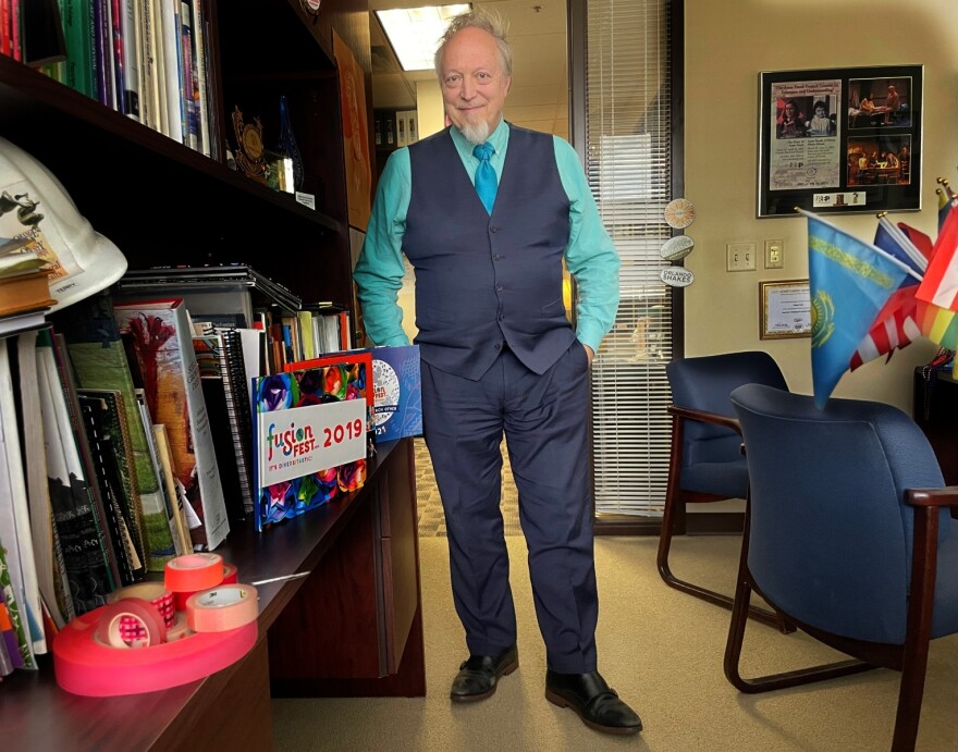 Terry Olson in his office at Orange County Arts and Cultural Affairs. Visible on the bottom left is what he calls his "cutting red tape sculpture." Small flags representing his multicultural works such as FusionFest and Diversitastic Dinners are seen on his desk at right.