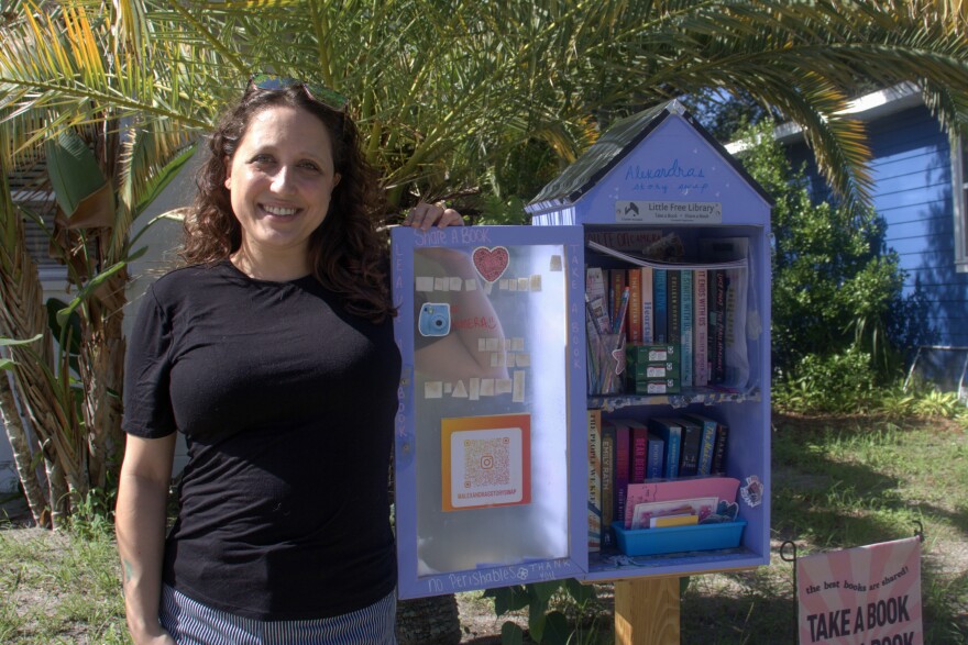 A smiling woman wearing a black shirt stands next to a lavender case of books.
