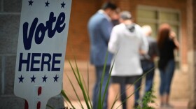 A "Vote Here" sign sits in the foreground. Five people line up in background waiting to vote.