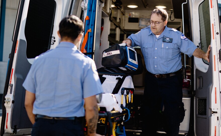 Two paramedics wearing light blue shirts remove a gurney from an ambulance. 