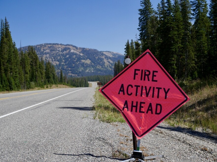 A red diamond-shaped road sign with the words “Fire Activity Ahead,” next to a two-lane highway with mountains and pine trees in the background.