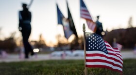 A small American flag pokes out of the ground, with other flags and two marching people in the background. 