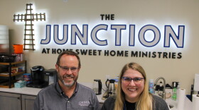 Home Sweet Home Ministries CEO Matt Burgess and Residential Manager Jenny Sharp stand behind a counter in the Junction. There are kettles and coffee makers behind them on the counter. Above and behind them is a sign for the Junction.