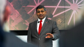 A man in a suit and tie with an OSU pin speaks on a stage. 