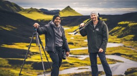 Two men pose with camera gear in a mountain landscape.