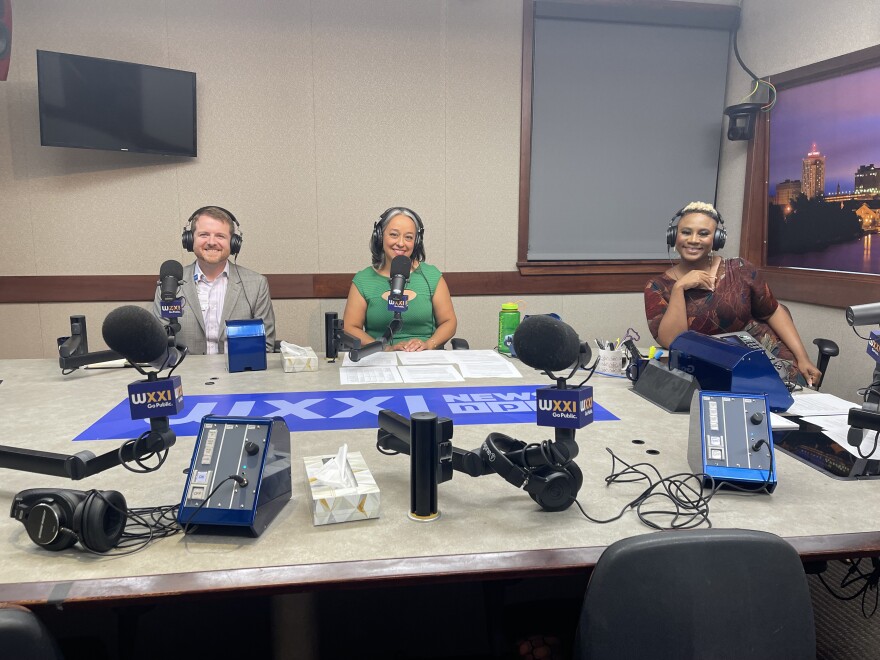 Three people wearing headphones sit at a table in a radio talk studio: a man at left has short brown hair, a brown beard and is wearing a light grey blazer over a white button-down shirt; a woman at center has shoulder-length grey hair and is wearing a green dress; a woman at right has short blonde hair and is wearing a brown dress.