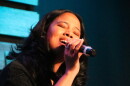  A woman sings into a handheld microphone with her eyes closed in a darkened performance space.