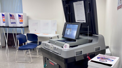 A gray voting machine sits open, with guarded places for voters to cast their ballots behind the machine.
