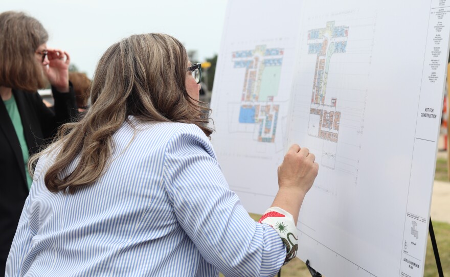 A woman in a striped shirt closely examines a detailed architectural blueprint displayed on an easel outdoors.