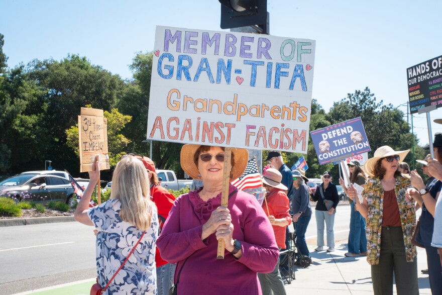 An older woman in a hat and sunglasses stands in a protest on a street corner. She holds a sign that reads, "member of gran-tifa: grandparent's against fascism."