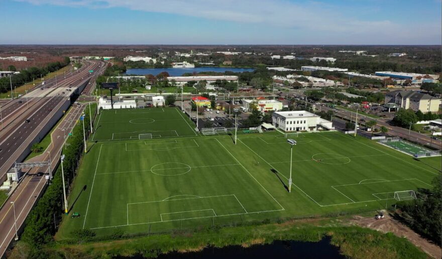 An overhead view of the Tampa Bay Rowdies training facility. Three soccer field are to the right of a road. There is a white building in front of one of the fields. There is a lake in the background.