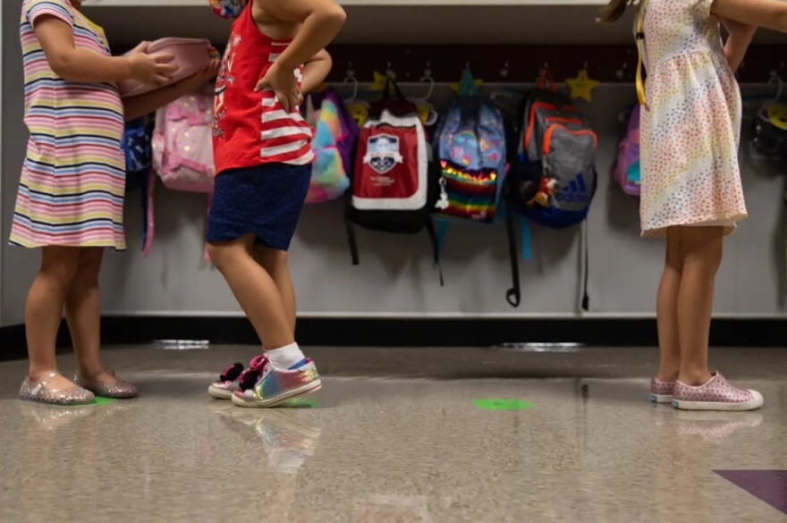 Kindergarten students are seen lined up in front of a backpack station, where all the bags are lined up and hanging on individual hooks.