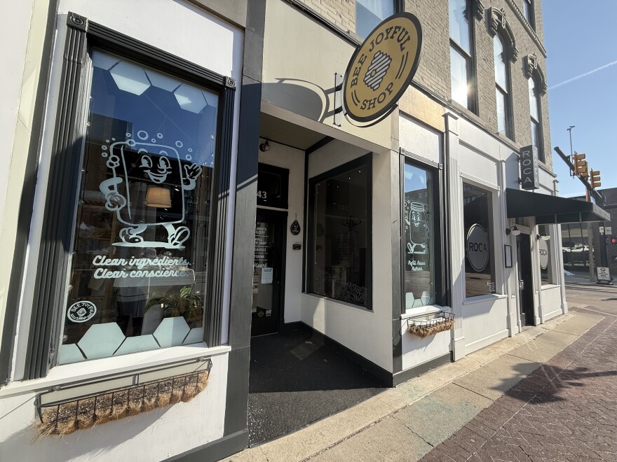 The front of Bee Joyful shop is depicted on the Kalamazoo Mall. The sidewalk in front of the store is red brick, and the storefront has two large glass windows with murals. One mural is of a cartoon bar of soap with a face and hands that is waving. Underneath it are the words “clean conscience”. The Bee joyful shop sign is a yellow circle with a bee hive in the middle.