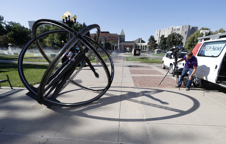 A bronze statue in a circular formation lays on the ground sidewise. A person from a news crew is filming it with a camera. 