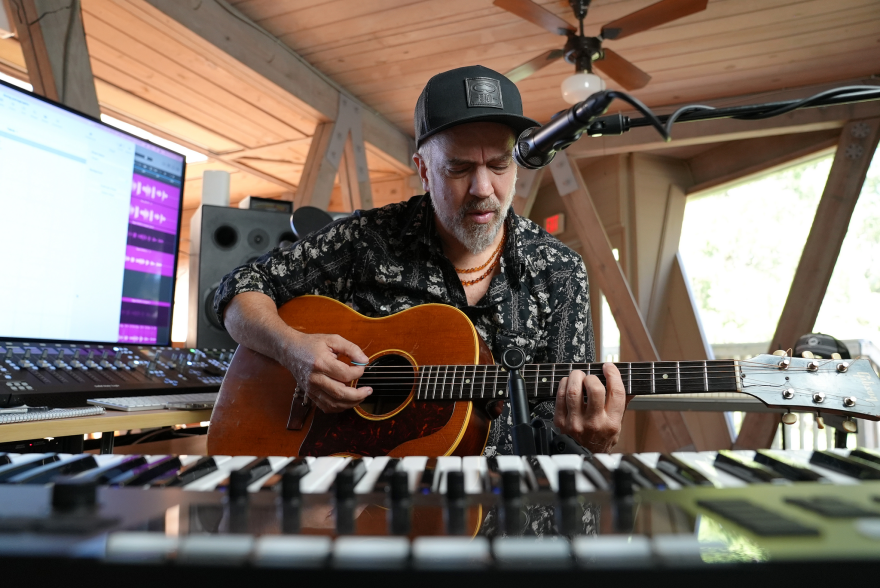 Surrounded by a keyboard and recording equipment, a musician sings and plays guitar.