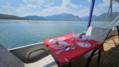 a table set for two on the deck of a boat has a red tablecloth, two beige and brown chairs. It's overlooking the water of Lake Dillon with mountains in the background