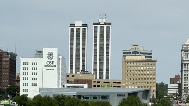 A skyline view of Peoria shows modern high-rise buildings, including two tall condominium towers, an OSF HealthCare facility, a tan historic-looking building, and trees in the foreground under a partly cloudy sky.