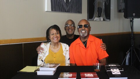 Three people sit smiling at a table covered with copies of a paperback book. A woman in a yellow top and white floral cardigan sits on the left, a man in a black shirt and glasses stands behind, and an older man in a bright orange shirt and sunglasses sits on the right.