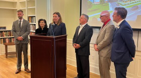 A woman is standing at a lectern speaking with a bunch of people in suits behind her. Everyone is in a library. There's a rendering of the project being projected onto a screen. 