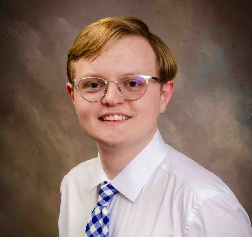 Nicholas Hazelett. A young, red-haired man wearing glasses and dressed in a white dress shirt and blue checkered tie.