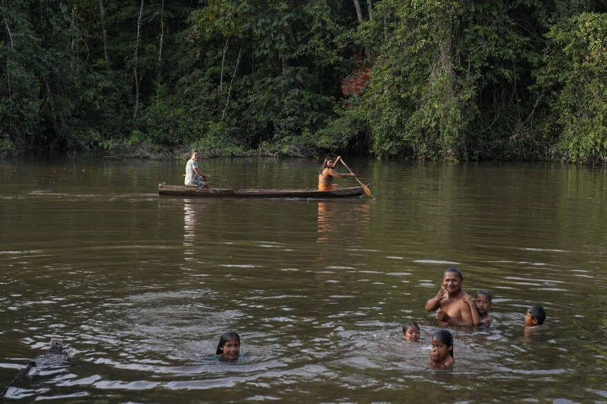 A man and five children play in the waters of a river, while a two women ride a canoe behind them.