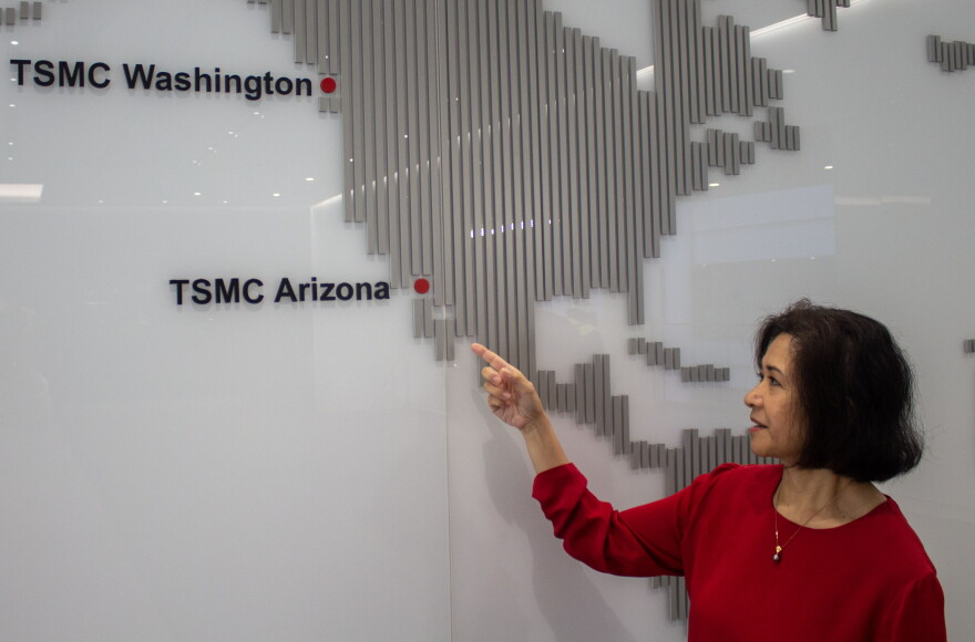 A woman in a red shirt points to a white and gray map that has a red dot where Arizona would be, and has in black block letters "TSMC Arizona." Above it, where Washington would be, is in big block letters "TSMC Washington" with another red dot representing the company's campus there. 