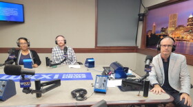Three people wearing headphones sit at a table in a radio talk studio: A woman back left has her dark hair pulled back. She is wearing glasses and a dark top under a black sweater with a white border; a man back center has short red hair and is wearing glasses and a blue and white plaid button-down shirt; a man front right has very short dark hair and is wearing glasses, a black button-down shirt under a grey suitcoat, jeans, and brown shoes.