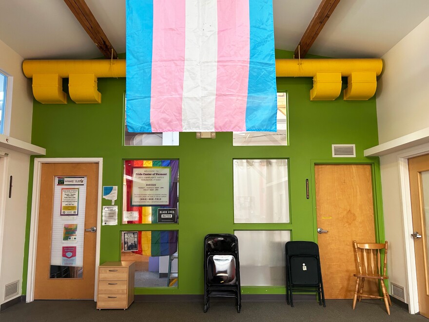 The trans pride flag, a flag with blue, pink and white stripes, hangs from the center of the ceiling in a room painted bright green with two wooden doors and some chairs.