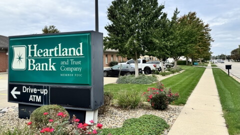 A Heartland Bank and Trust sign directs customers toward the ATM in front of a row of trees.