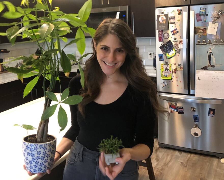 A woman smiles while she holds a tall plant with a twisted trunk and sets of five leaves in one hand and the other holds a small plant with spiky leaves, similar to a coral reef. She’s standing inside a kitchen.